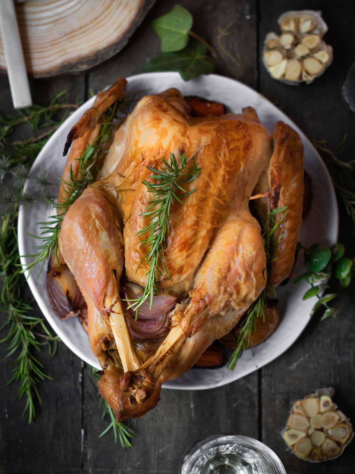 Roasted turkey on a plate, garnished with fresh rosemary, surrounded by garlic cloves and a glass of water on a dark wooden table.