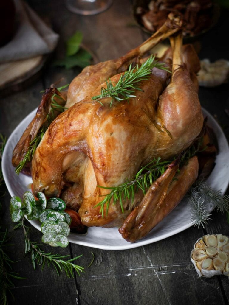 Roasted turkey garnished with rosemary on a white plate, surrounded by herbs and a halved roasted garlic bulb.