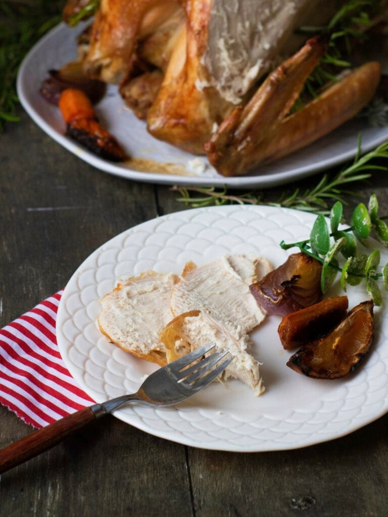 Sliced roasted turkey breast with herbs and roasted onions on a white plate, featuring a red-striped napkin and fork.