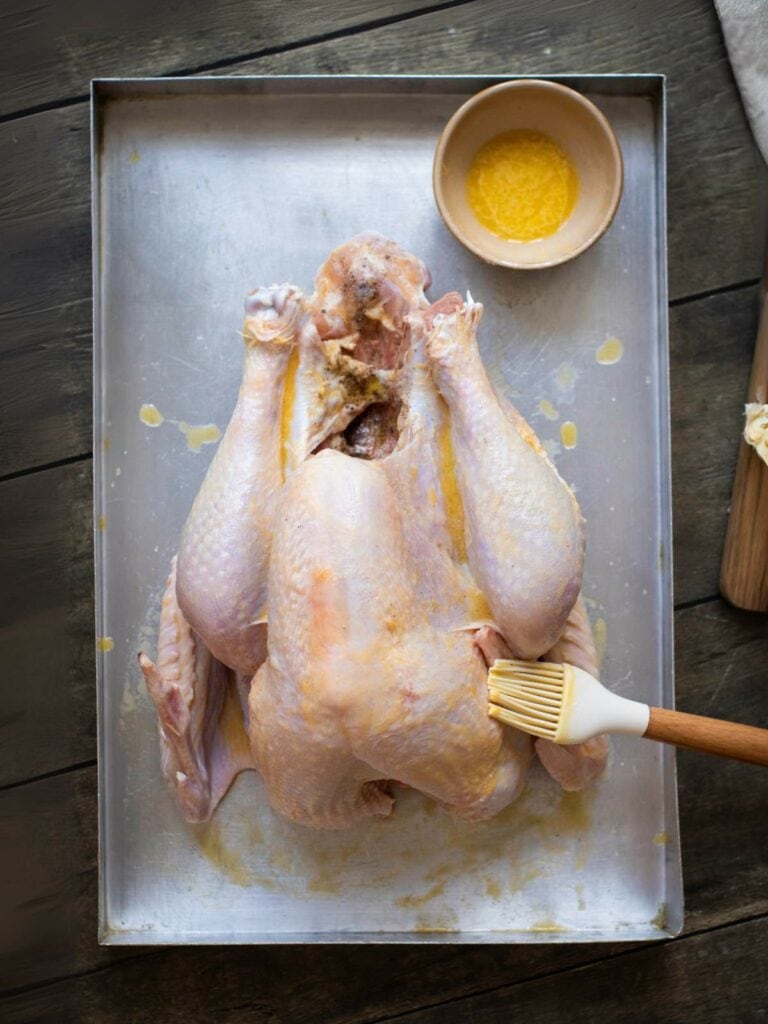 Raw turkey on a metal baking tray being brushed with melted butter from a bowl next to it.