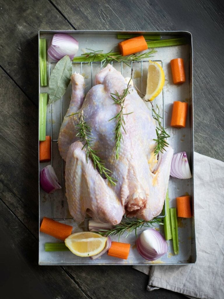 Raw whole chicken on a tray with rosemary, lemon wedges, carrot pieces, celery, red onion, and bay leaves on a wooden surface.