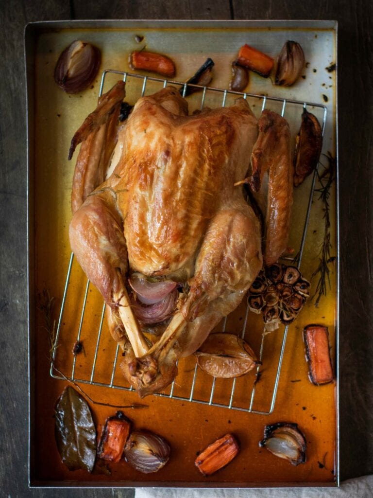 Roasted chicken on a rack in a baking tray, surrounded by roasted garlic, carrots, onions, and herbs.