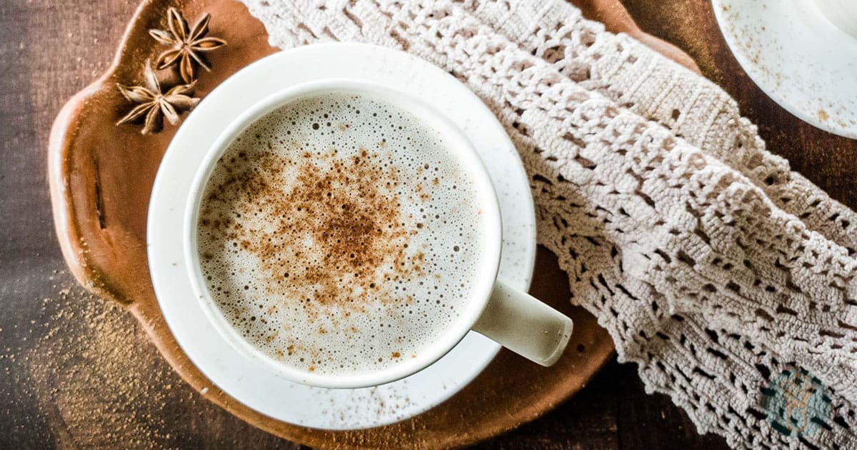 A cup of chocolate chai tea with a sprinkle of spice sits on a wooden tray, accompanied by star anise and a decorative lace cloth.