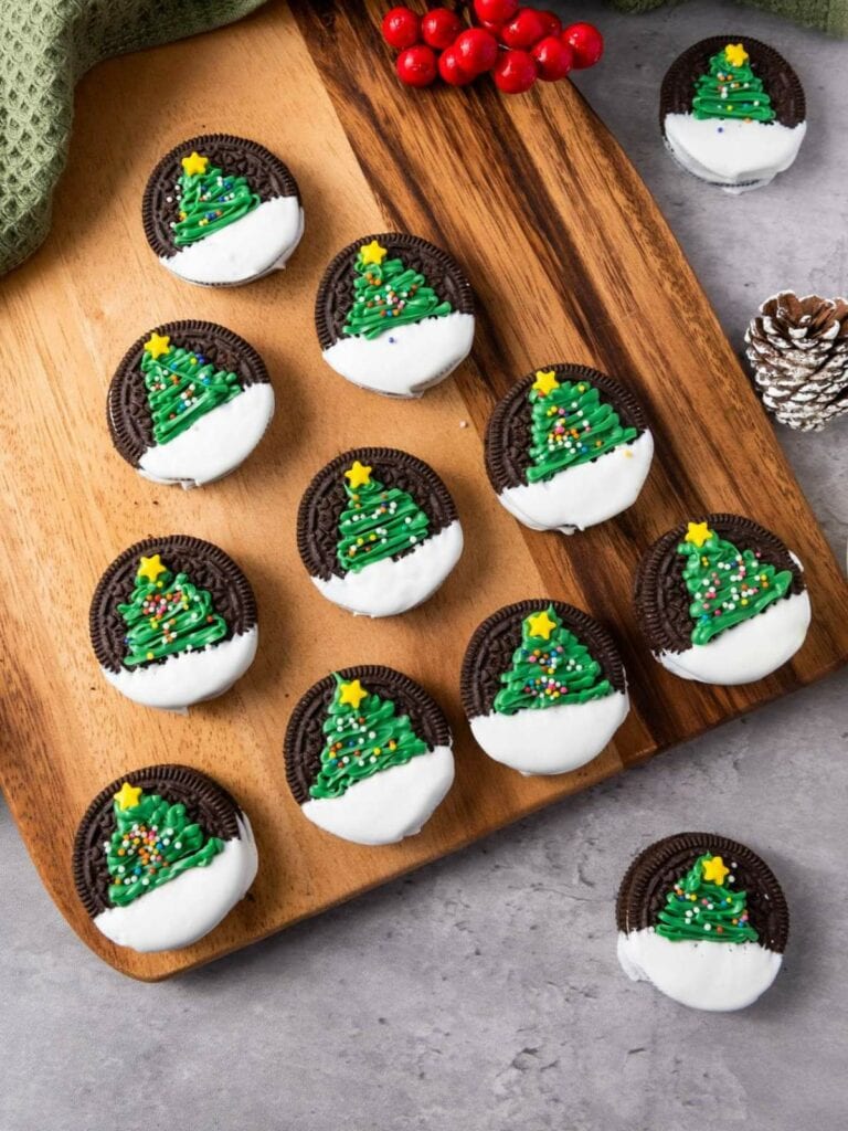 Christmas tree oreos arranged on a wooden board. Red berries and a pine cone are nearby.