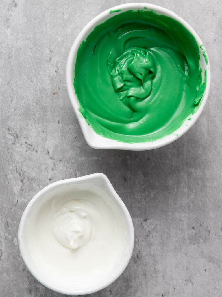 Two bowls of frosting on a gray surface; one with green frosting and one with white frosting.