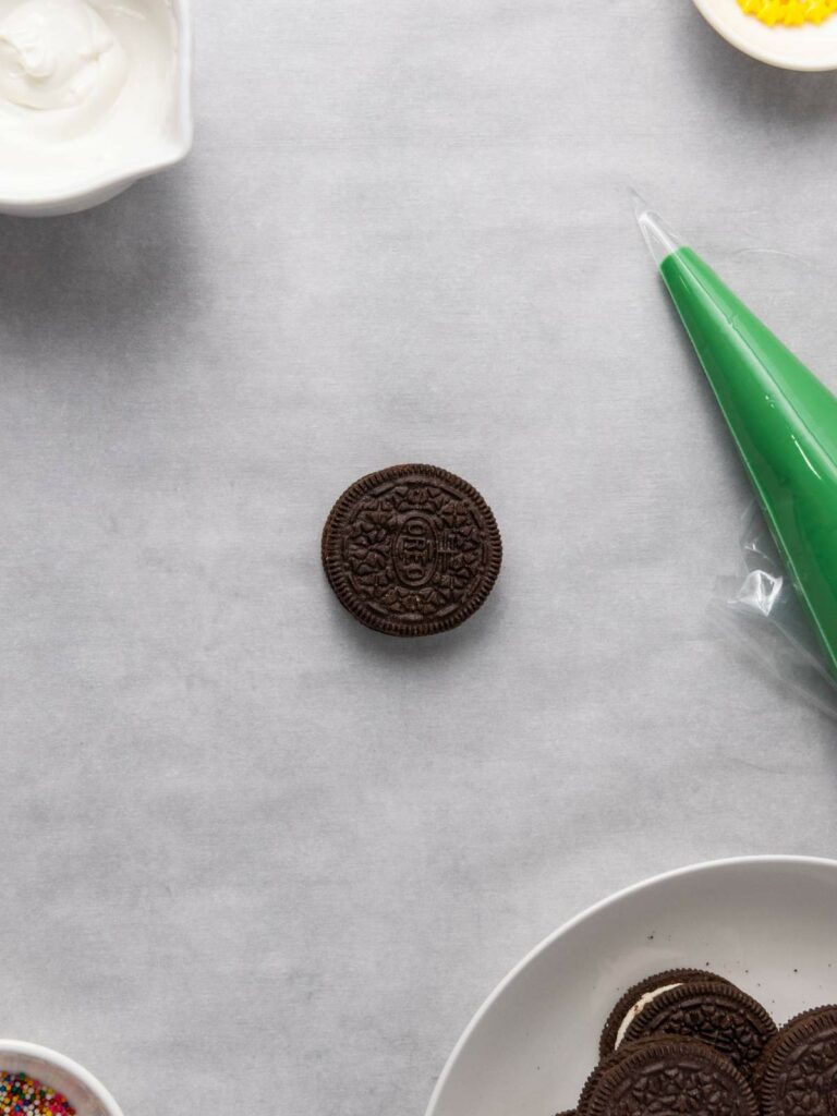 A single chocolate sandwich cookie is placed on a gray surface, surrounded by icing and small bowls of frosting and sprinkles.