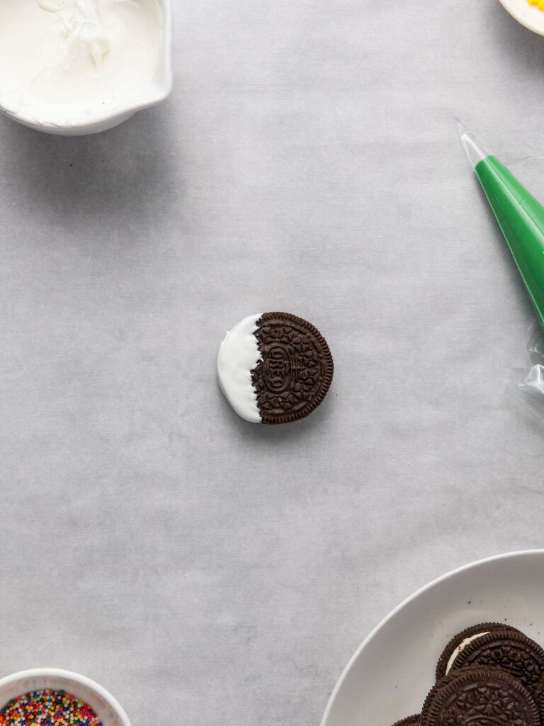 Chocolate cookie partially dipped in white frosting on a parchment paper surface, surrounded by a bowl of frosting, a piping bag, and a plate with similar cookies.