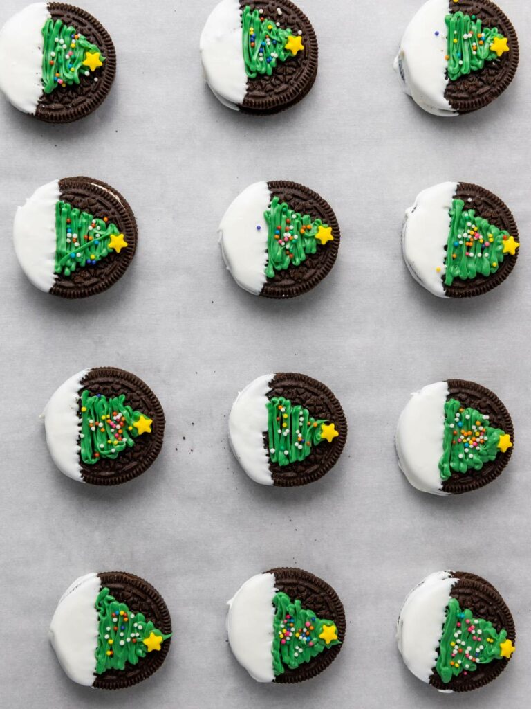 Twelve chocolate sandwich cookies decorated with green Christmas trees and star sprinkles, partially dipped in white coating.