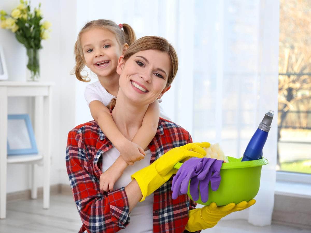 A smiling woman holding cleaning supplies with a child hugging her from behind in a bright room.