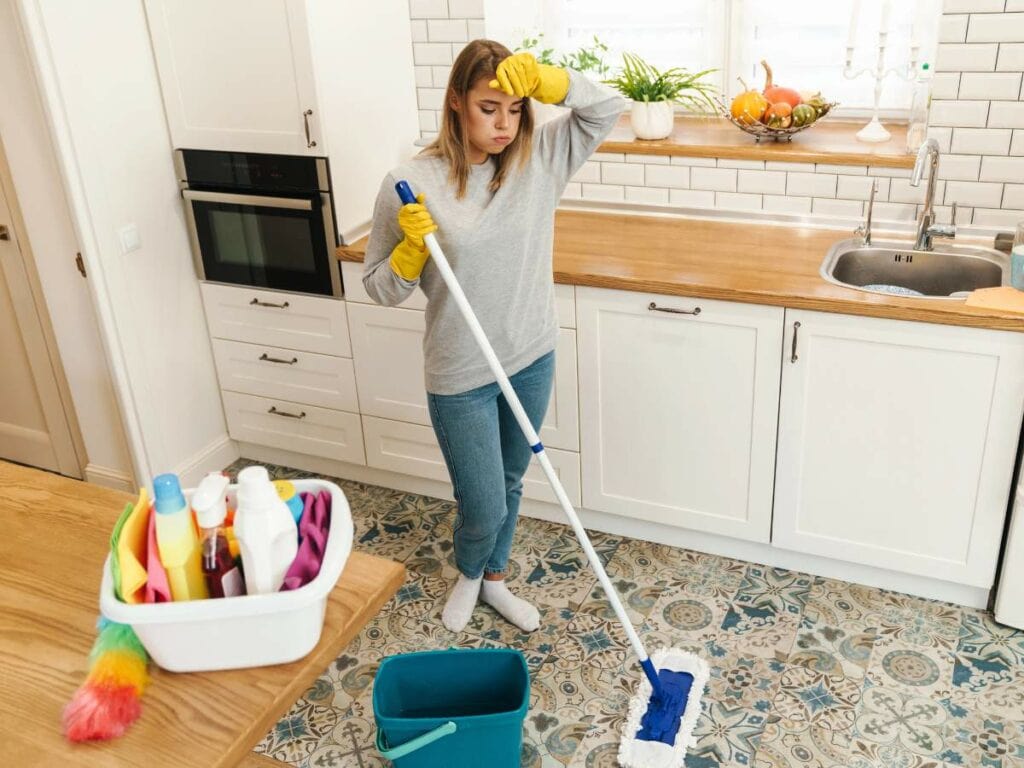 A woman in a kitchen pauses while mopping the floor, with cleaning supplies on the counter and a mop bucket nearby.