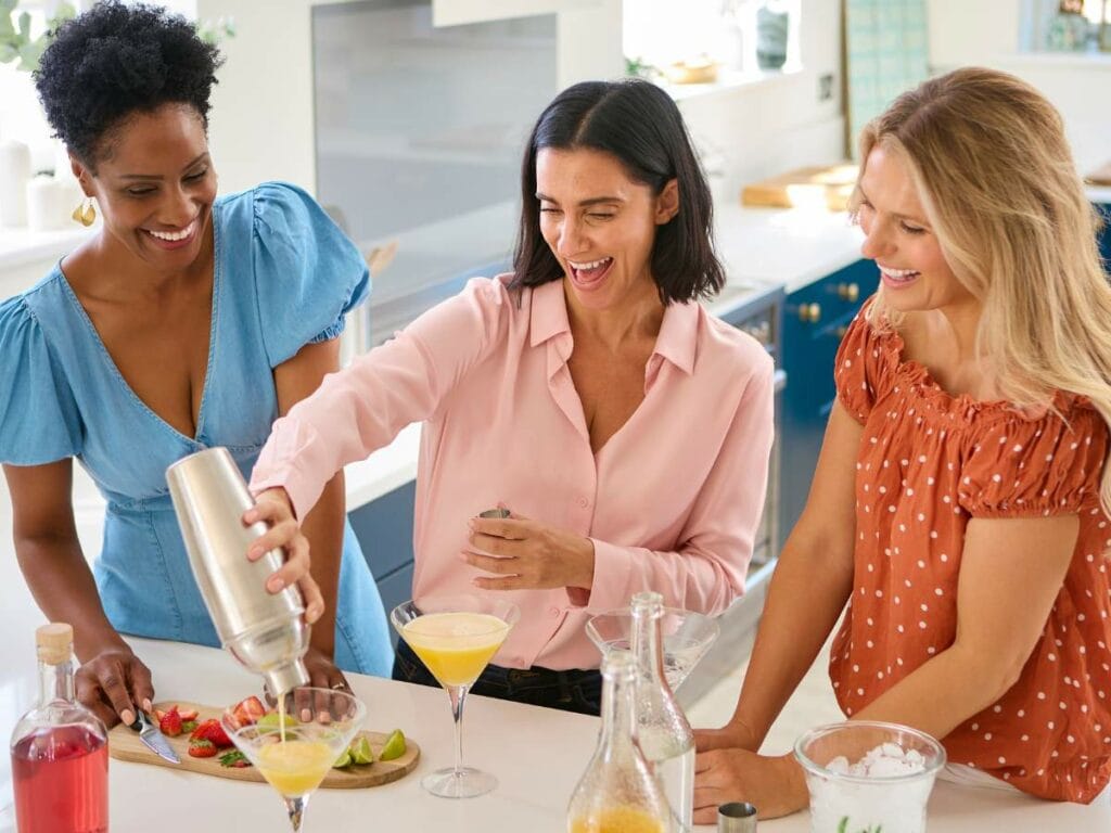 Three women in a kitchen making cocktails. One is using a shaker while the others smile. Ingredients and drinks are on the counter.