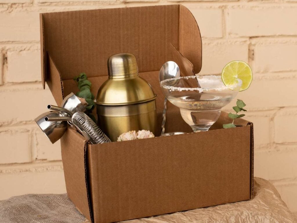 Cardboard box containing a cocktail shaker, jigger, strainer, margarita glass with lime, and a cork.