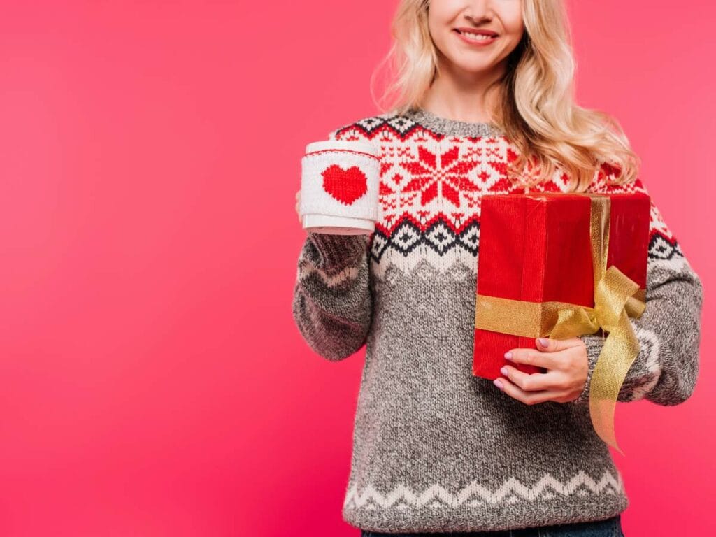 Person in a patterned sweater holding a mug with a heart design and a red gift box.