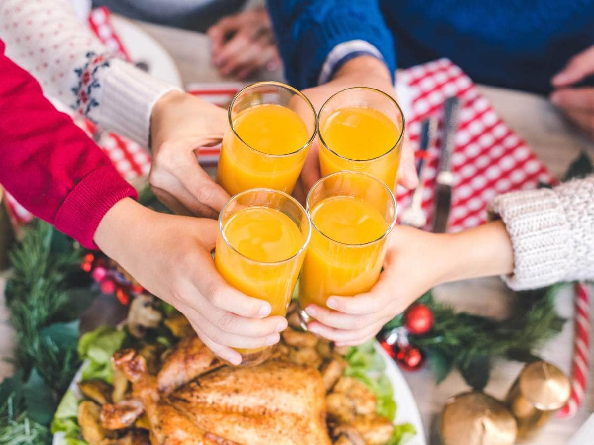 People holding glasses of orange juice over a table set with a roasted chicken and festive decor.