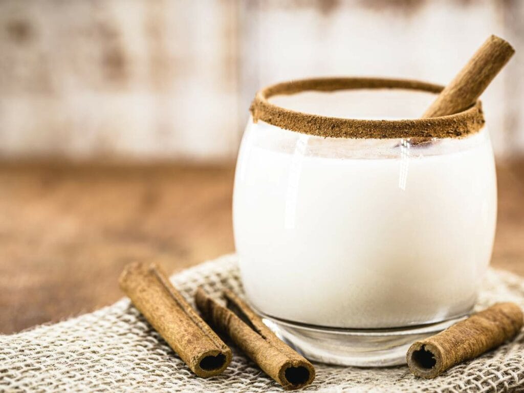 A glass of milk with a cinnamon stick and rim sits on a woven placemat.