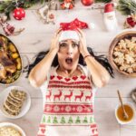 A woman in a Santa hat looks surprised while lying on a kitchen floor surrounded by Christmas foods and decorations.