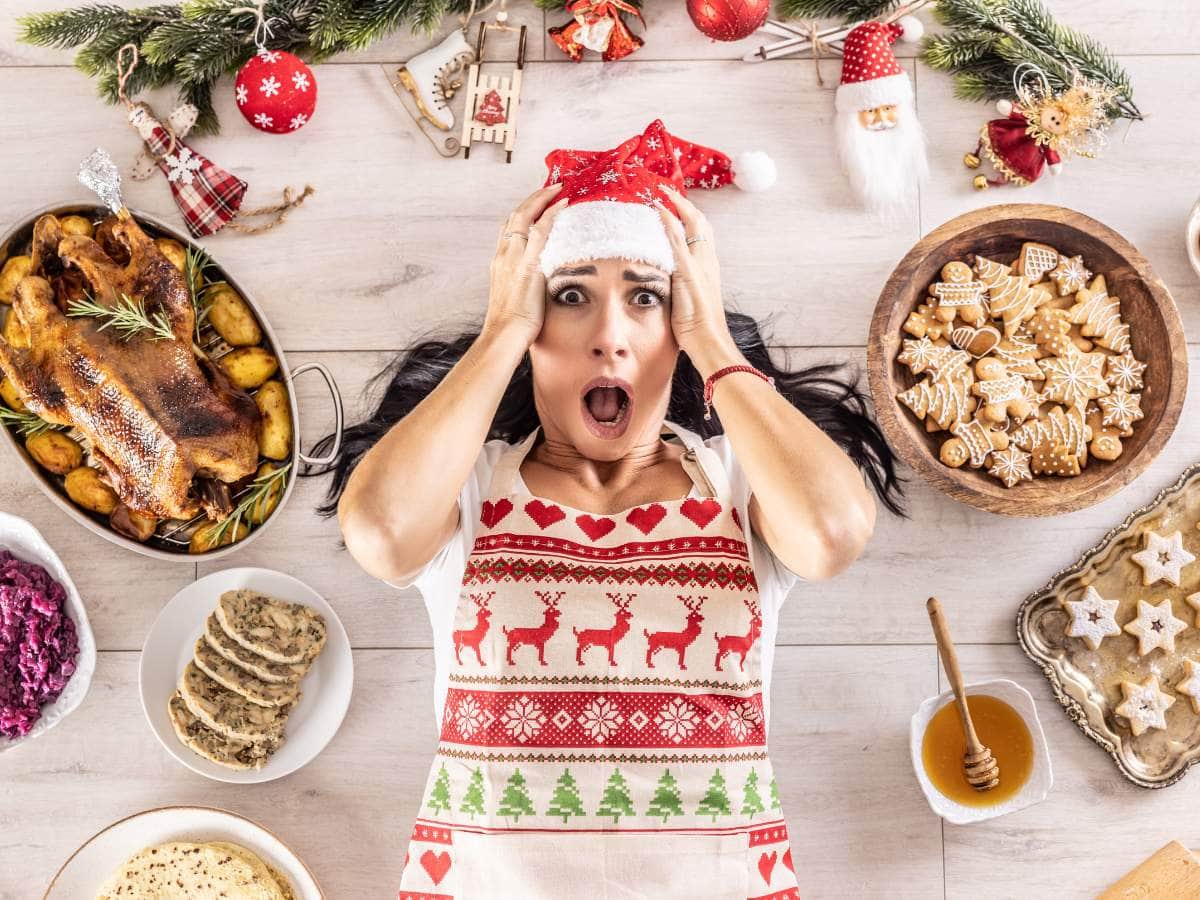 A woman in a Santa hat looks surprised while lying on a kitchen floor surrounded by Christmas foods and decorations.