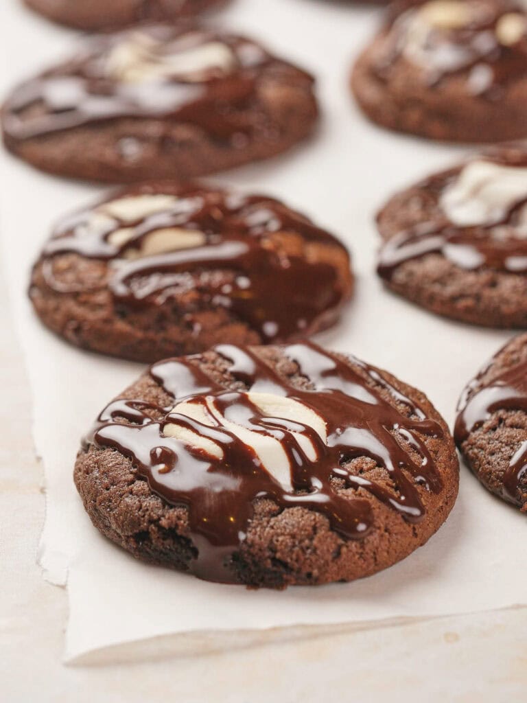 Chocolate cookies with white chocolate chunks, drizzled with dark chocolate, on parchment paper.