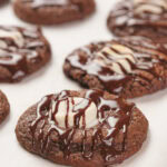 Chocolate cookies with white chocolate centers and drizzled chocolate on top, arranged on parchment paper.