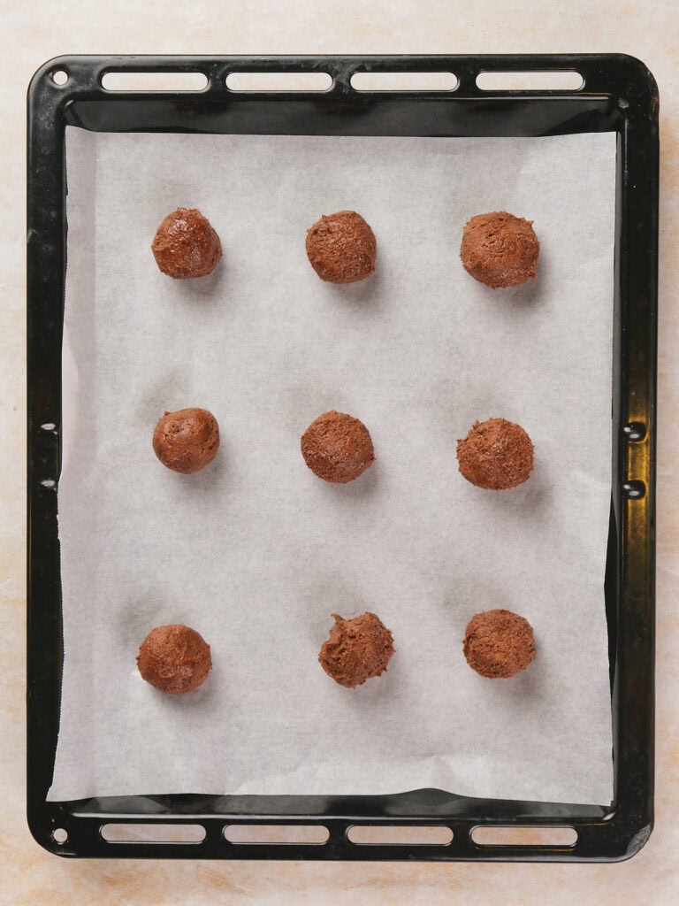 Baking tray with nine chocolate cookie dough balls on parchment paper.