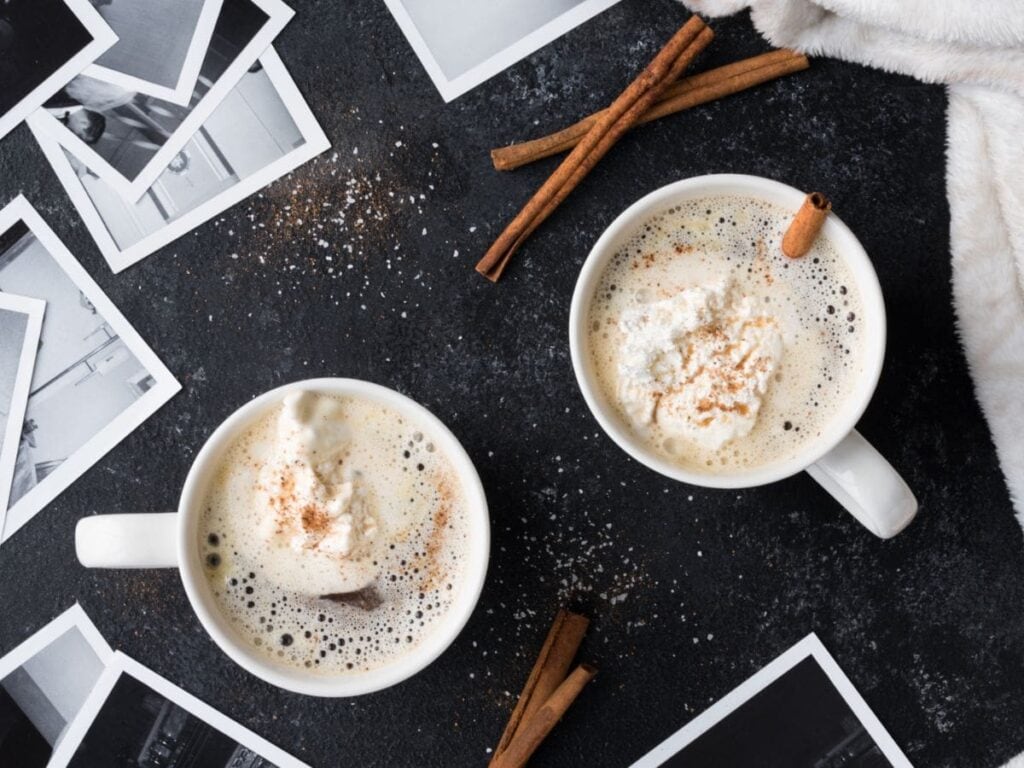 Two mugs of frothy coffee topped with whipped cream and cinnamon on a dark surface. Surrounded by scattered black-and-white photos and cinnamon sticks.