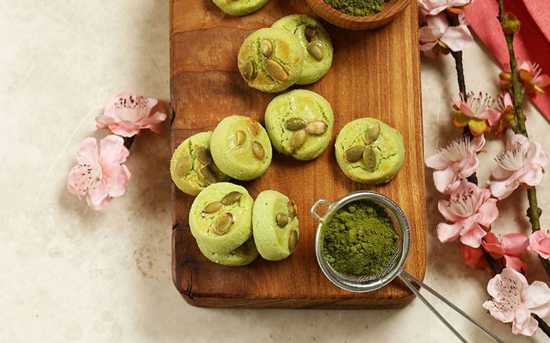 Matcha sugar cookies on a wooden board, surrounded by pink flowers and a small sieve with matcha powder.