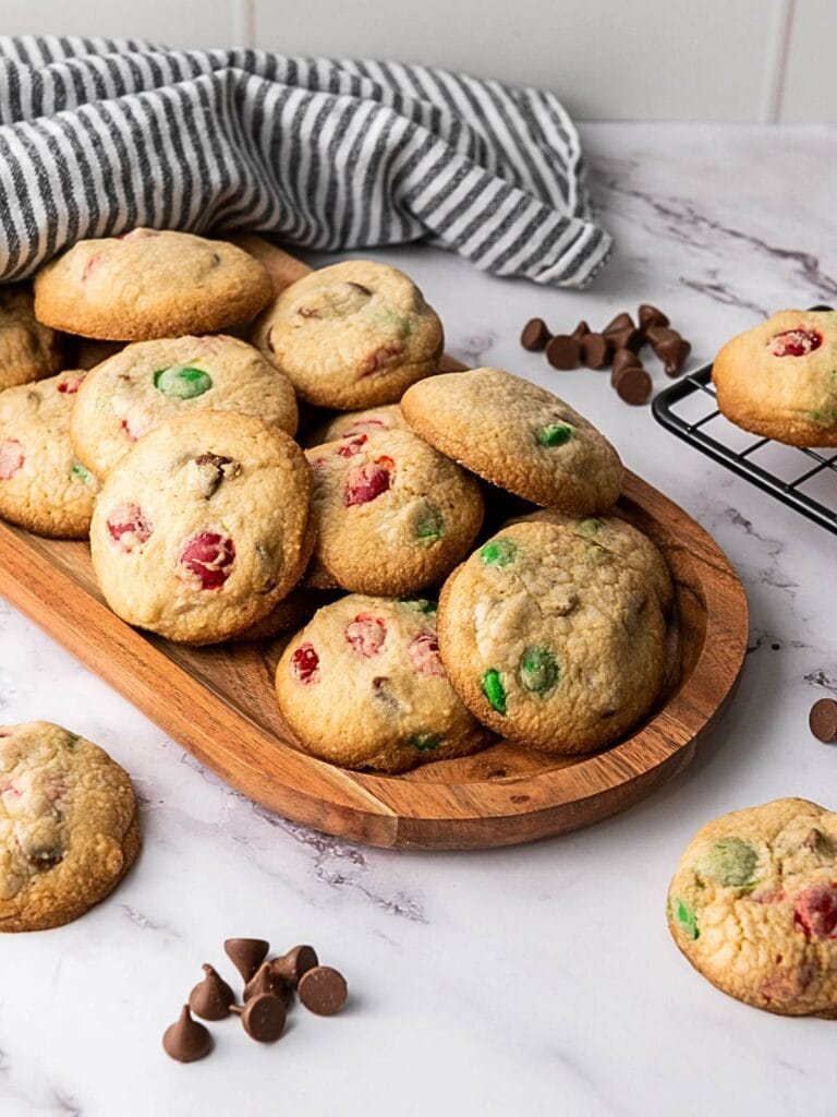 M&M cookies for Christmas arranged on a wooden board, with a striped cloth and chocolate chips.