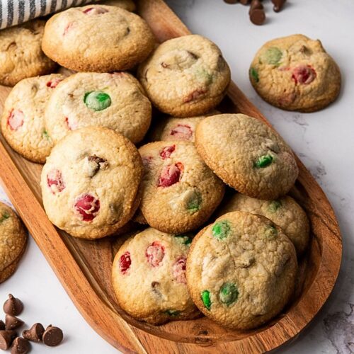 Wooden platter with M&M cookies for Christmas containing red and green candies and chocolate chips, placed on a marble surface with chocolate chips.