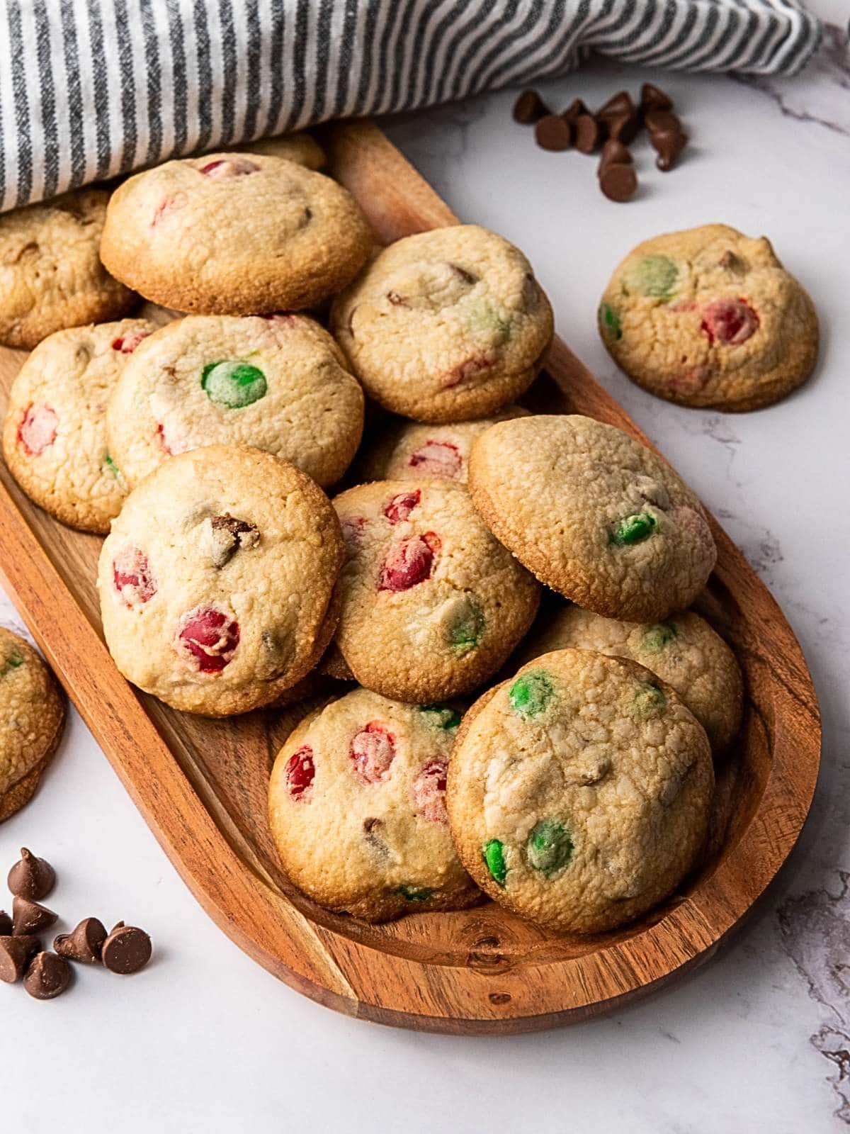 Wooden platter with M&M cookies for Christmas containing red and green candies and chocolate chips, placed on a marble surface with chocolate chips.