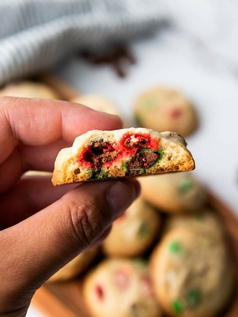 Close-up of a hand holding a half-eaten M&M cookies for Christmas with colorful candy pieces inside.