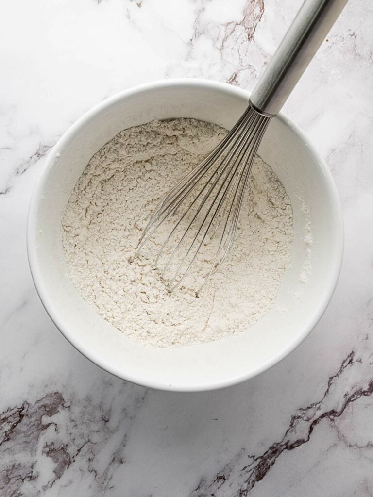 A metal whisk in a white bowl of flour on a marble surface.