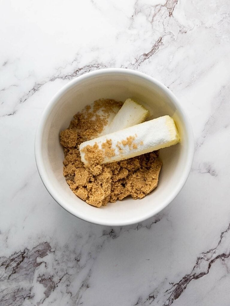 White bowl with brown sugar and two sticks of butter on a marble surface.