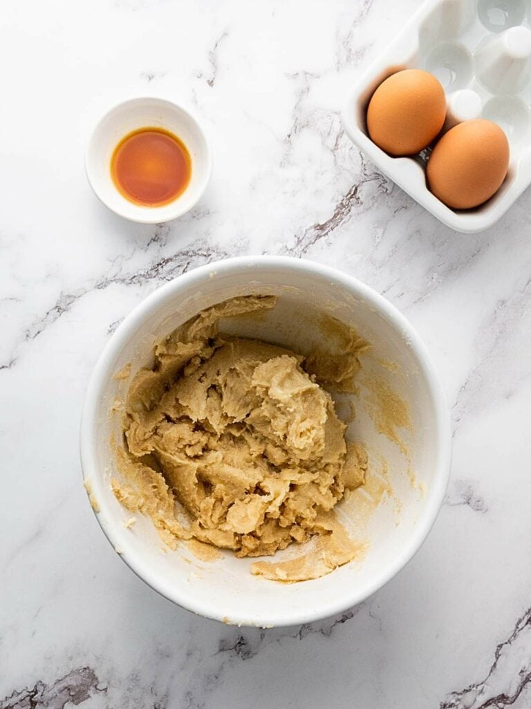 Bowl of cookie dough next to eggs and a small dish of vanilla extract on a marble surface.