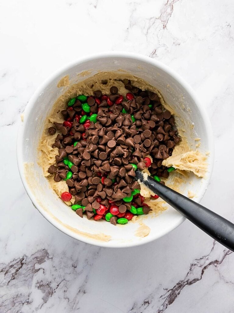 Bowl of cookie dough with chocolate chips and red and green candies, with a black spoon on a marble surface.