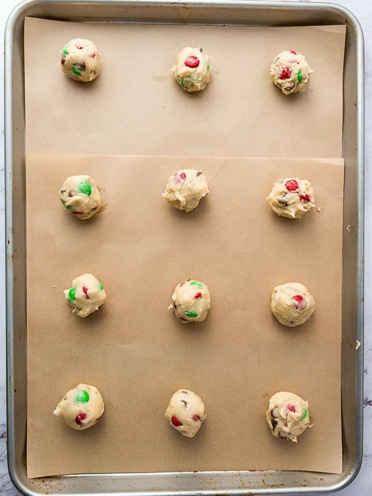 Cookie dough balls with red and green candies on a parchment-lined baking sheet, ready to bake.