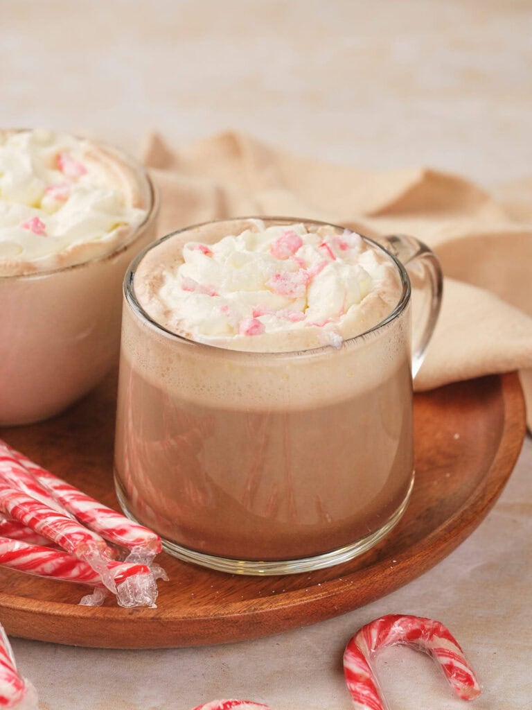 A glass mug of peppermint mocha topped with whipped cream and crushed peppermint rests on a wooden tray.