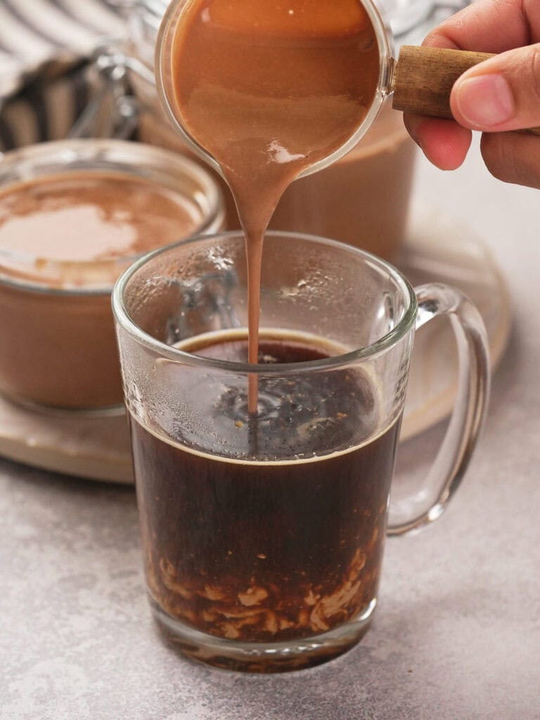 A hand pouring peppermint mocha coffee creamer into a clear mug of dark liquid.