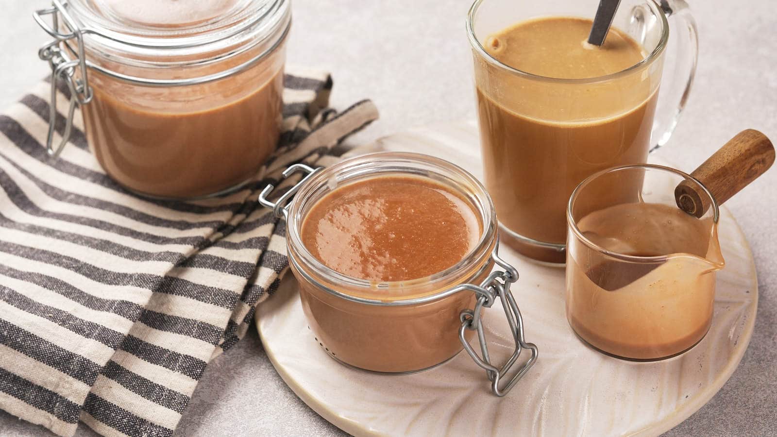 Jars and glass of brown liquid on a plate with a striped cloth nearby, including a glass container with a wooden-handled spoon.