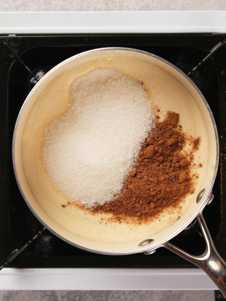 A saucepan on a stove contains cream with cocoa powder and sugar on top.