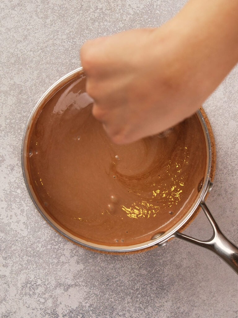 A hand stirs a chocolate mixture in a saucepan on a gray textured surface.