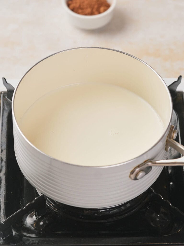 White pot filled with milk on a stovetop burner, with a small bowl of cocoa powder in the background.