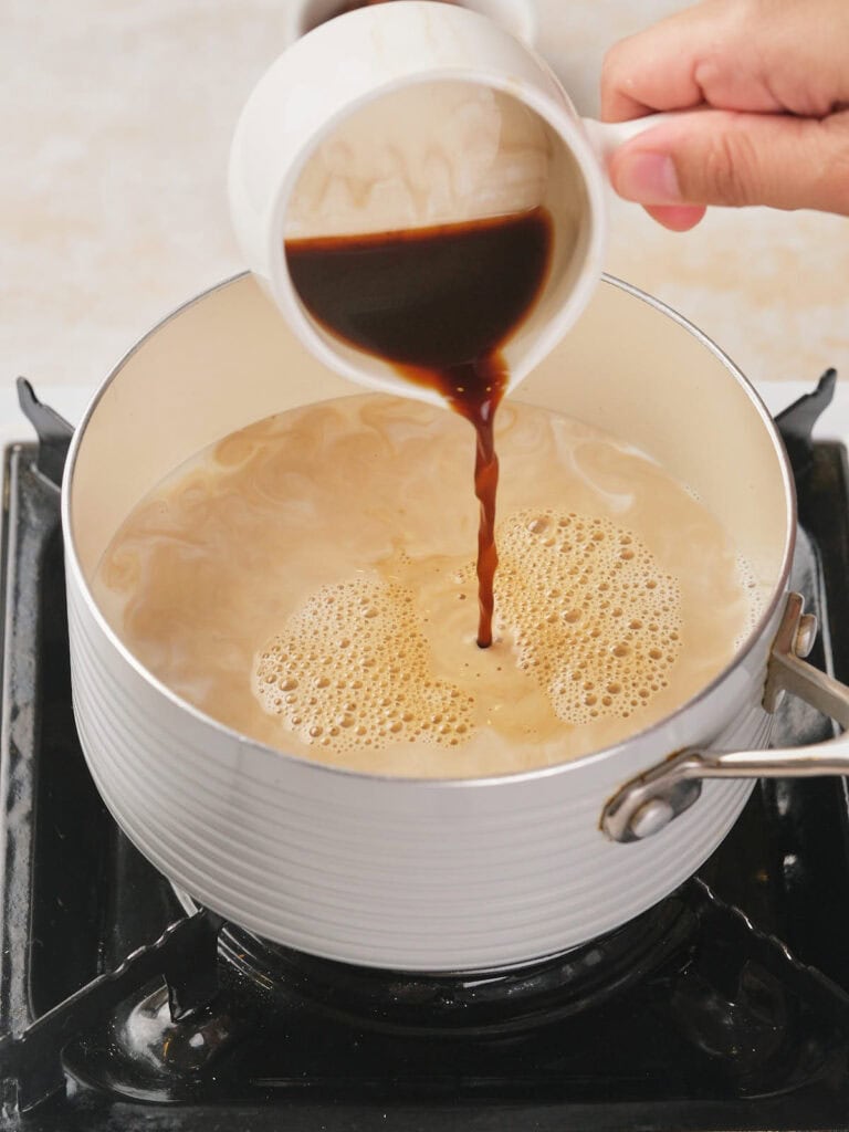 A hand pours coffee from a cup into a pot on a stove, creating a frothy mixture.