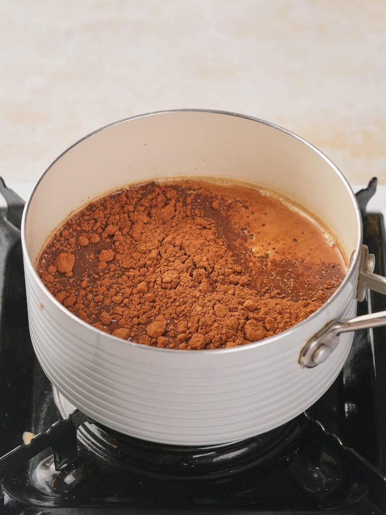 A white saucepan on a stove is filled with hot liquid and sprinkled with cocoa powder on top.