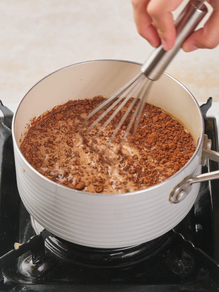 A hand is whisking cocoa powder and liquid in a white saucepan on a stovetop.