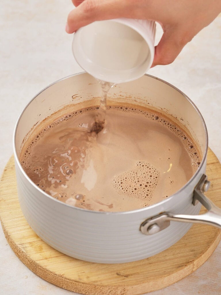 Hand pouring liquid into a saucepan containing frothy chocolate mixture.