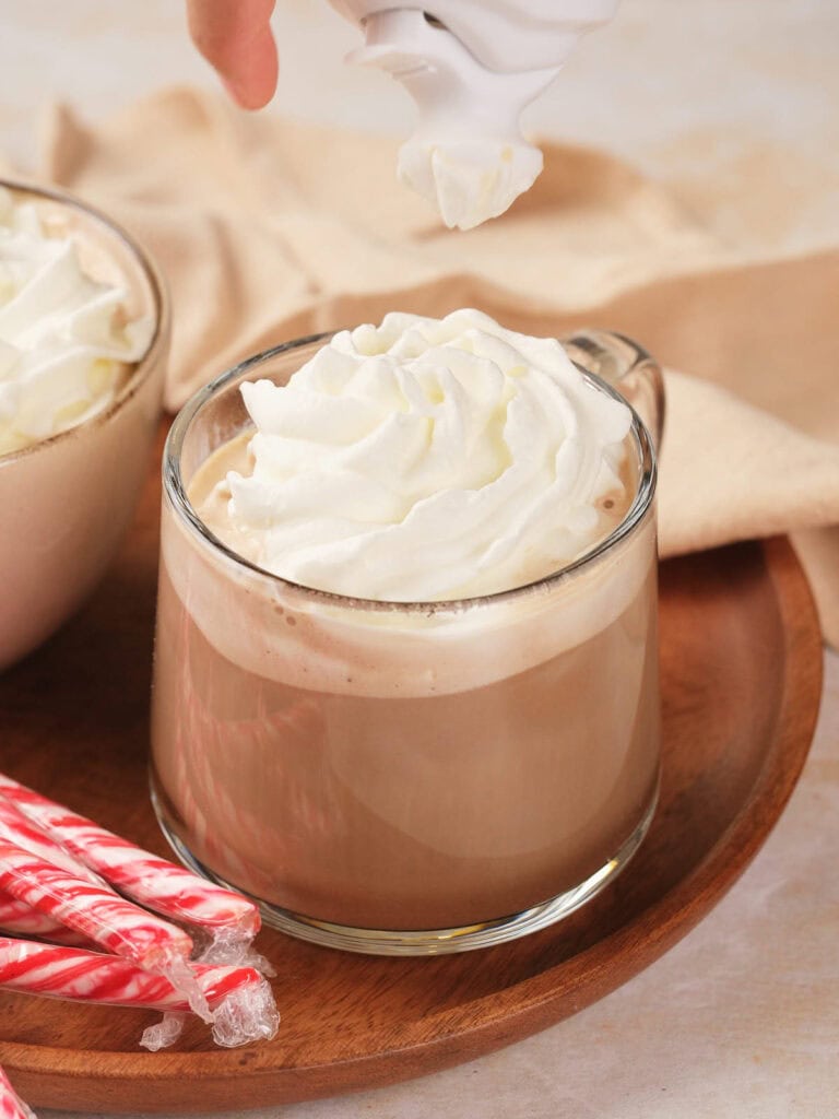 A glass mug of hot chocolate topped with whipped cream on a wooden tray.