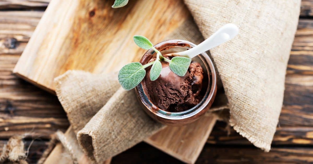 A jar of chocolate sage ice cream with a small green leaf garnish, placed on a rustic wooden surface with a burlap cloth underneath.