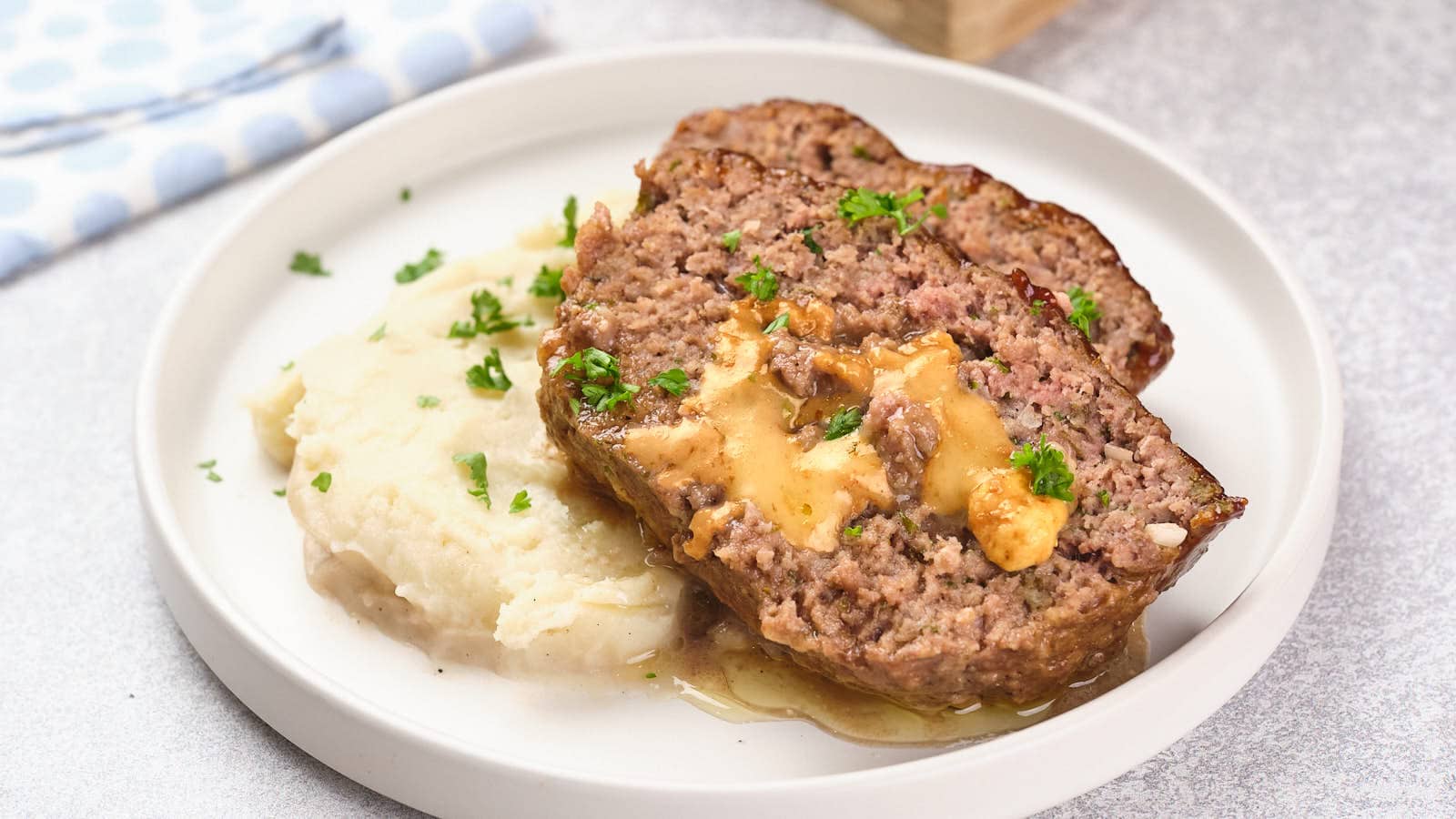 A plate with two slices of stuffed meatloaf topped with gravy, alongside mashed potatoes garnished with green herbs.