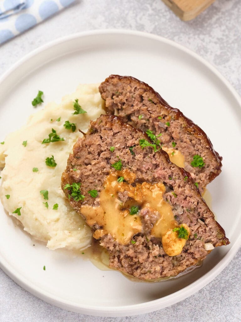 Two slices of stuffed meatloaf with melted cheese in the center served next to mashed potatoes, garnished with chopped parsley on a round white plate.