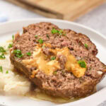 A slice of stuffed meatloaf with melted cheese on top, served on a white plate with mashed potatoes.